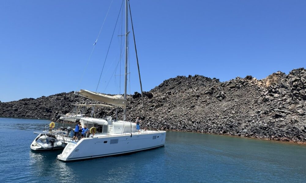 A white Santorini catamaran anchored in clear turquoise waters near volcanic cliffs on a sunny day.