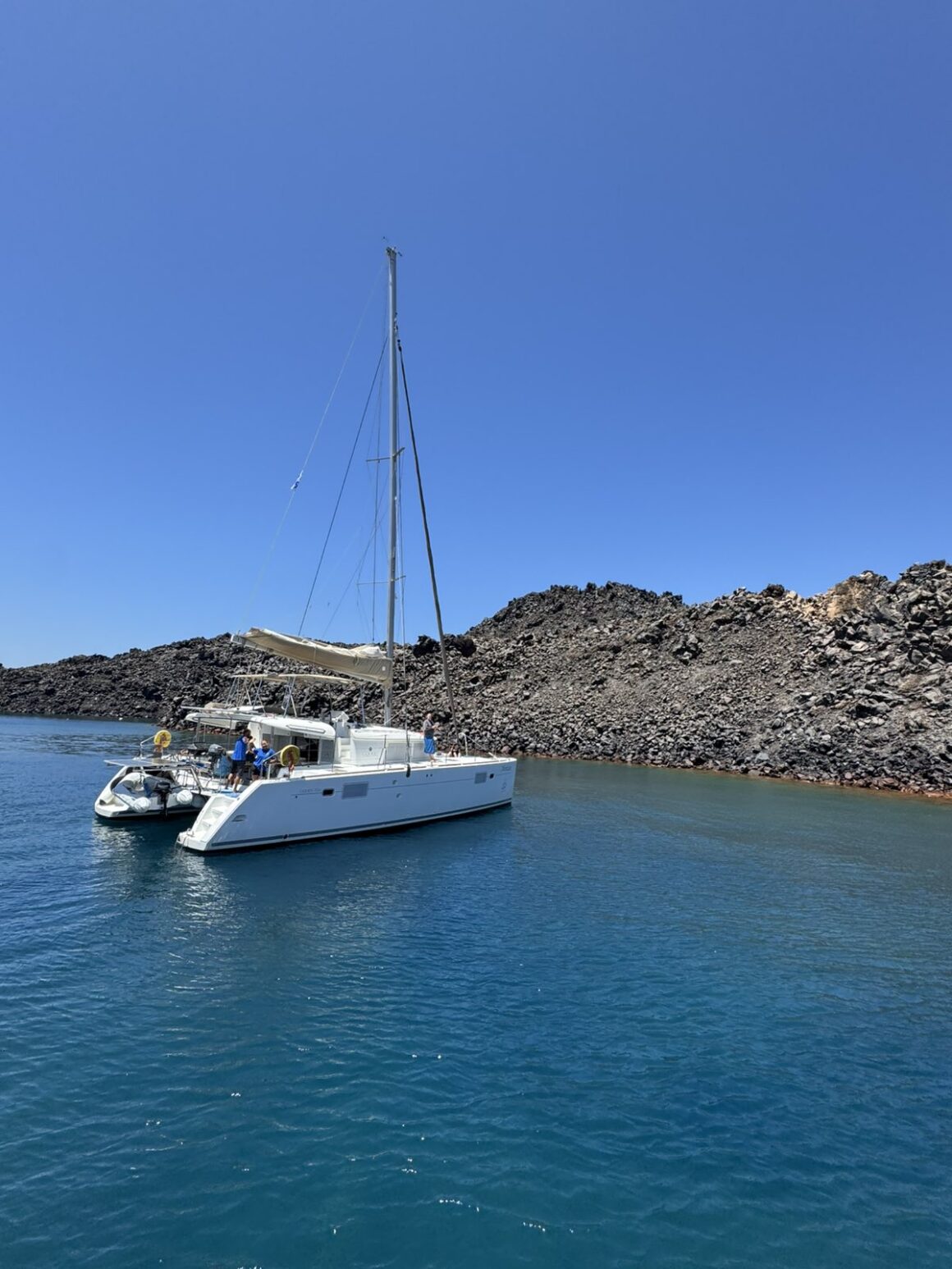 A white Santorini catamaran anchored in clear turquoise waters near volcanic cliffs on a sunny day.