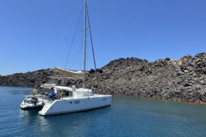 A white Santorini catamaran anchored in clear turquoise waters near volcanic cliffs on a sunny day.