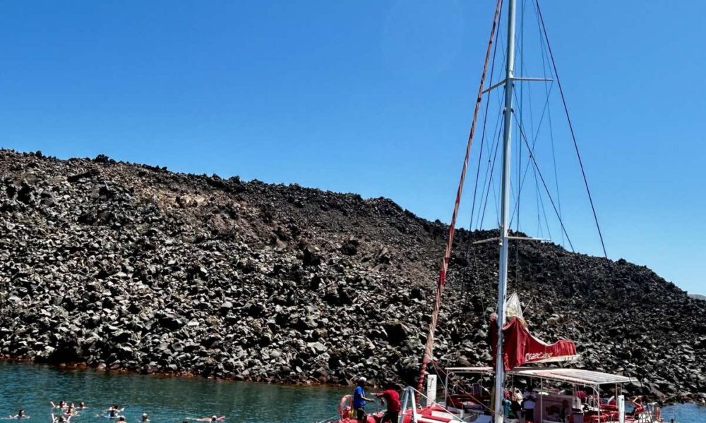 A red catamaran from Santorini Sailing anchored near the volcanic coast of Santorini under a clear blue sky.