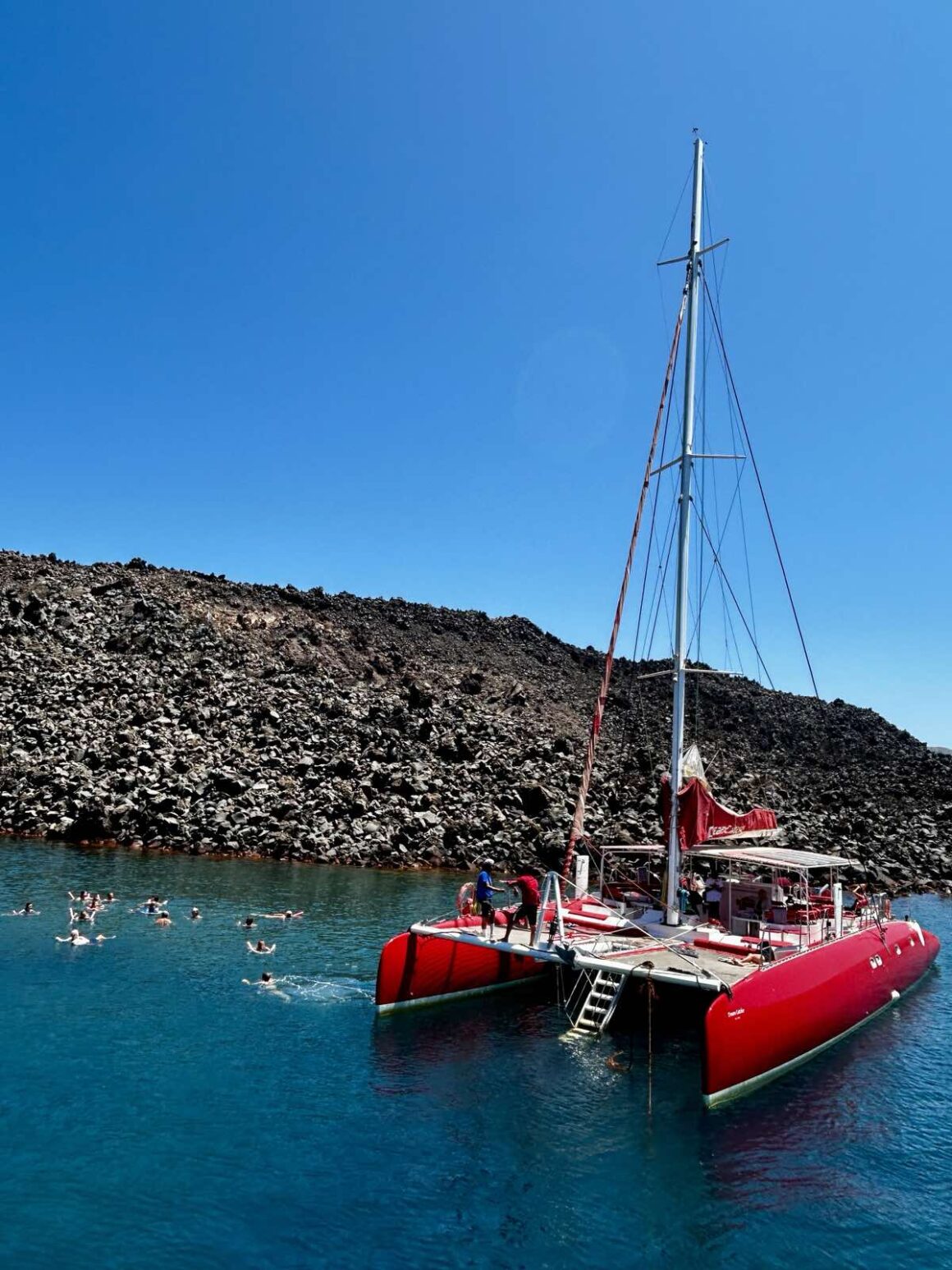 A red catamaran from Santorini Sailing anchored near the volcanic coast of Santorini under a clear blue sky.